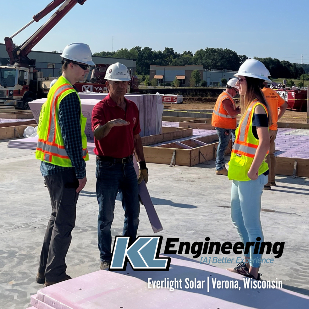 Two men and one woman, all wearing hard hats and safety vests, talking. In the background is a construction crane and cement work activities. KL Engineering logo.