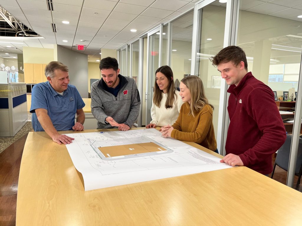 Five staff in a standing meeting in discussion, looking at plans on a table.