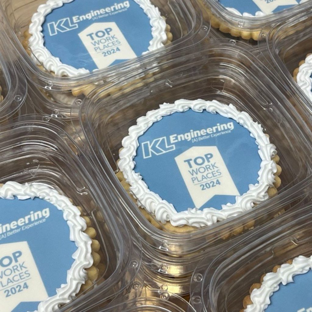 Photo of cookies with light blue frosting background with white KL Engineering logo over a white Top Workplaces logo with a white scalloped edge of frosting. The cookies are individually packaged and stacked in clear snap-top containers.