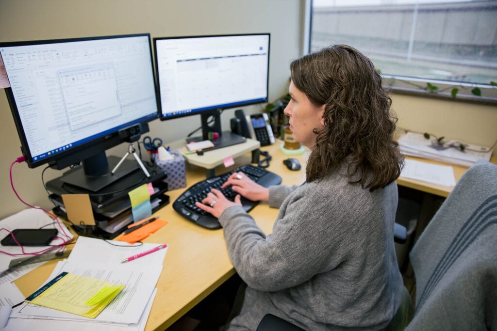 Photo of woman working at a computer with two monitors near a window.