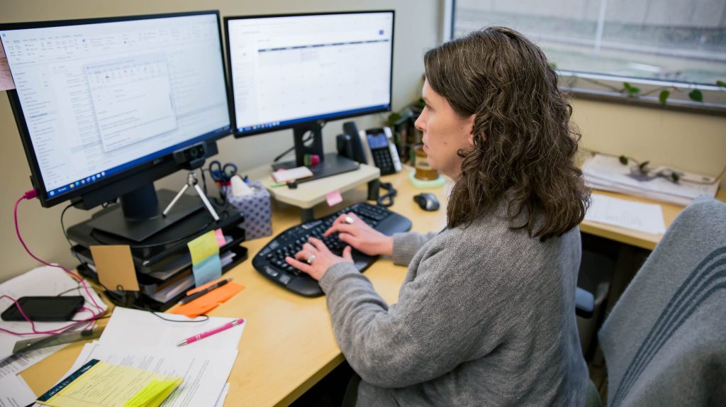 Photo of woman working at a computer with two monitors near a window.